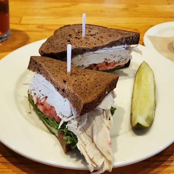 A turkey sandwich on dark bread with lettuce and tomato, served with a pickle spear.