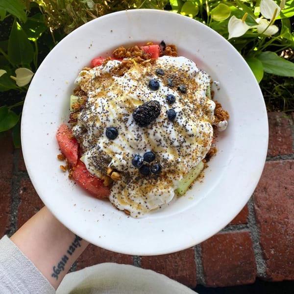 A white bowl of fruit, yogurt, granola, and berries, with a brick background.