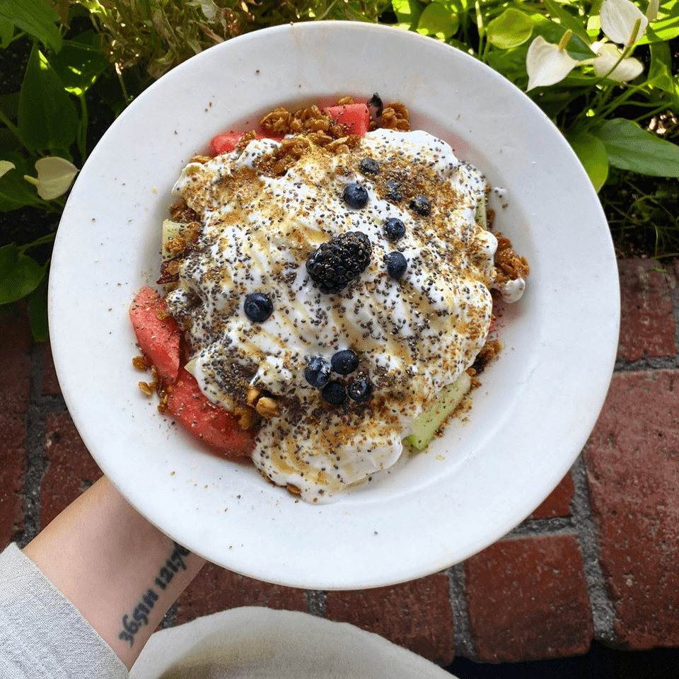 A white bowl of fruit, yogurt, granola, and berries, with a brick background.
