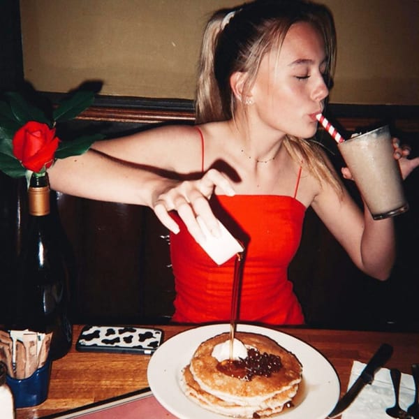 A young woman in a red top pours syrup on pancakes and sips a milkshake at The Kettle diner.