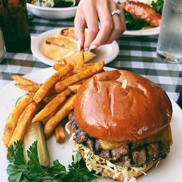 A diner burger with fries, pickle, and parsley on a white plate, with a hand reaching for a fry.