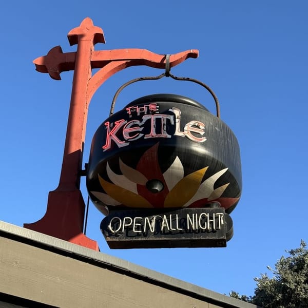 The Kettle diner's sign, shaped like a black kettle with "OPEN ALL NIGHT" in neon, against a blue sky.