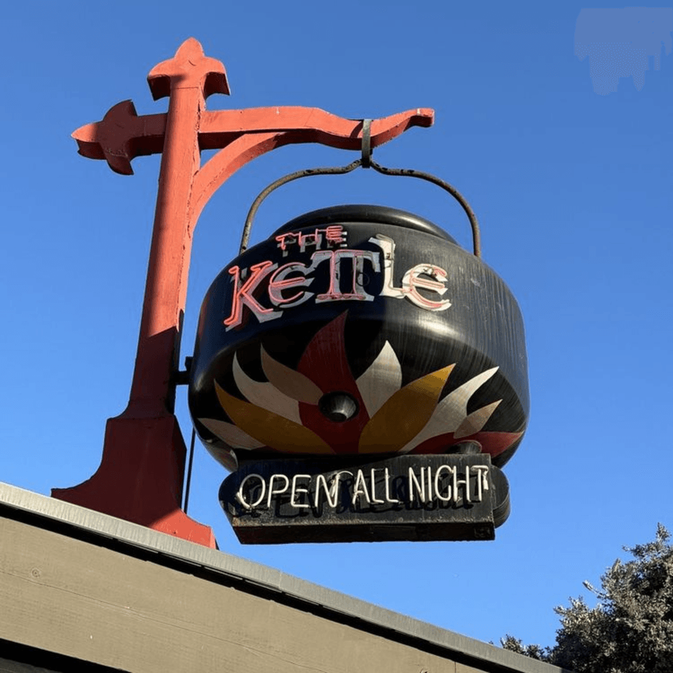 The Kettle diner's sign, shaped like a black kettle with "OPEN ALL NIGHT" in neon, against a blue sky.