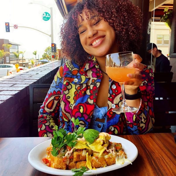 Smiling woman in a colorful jacket holding a mimosa, with a plate of tater tot nachos.
