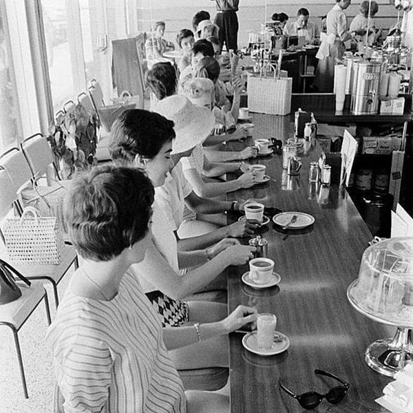 Black and white photo of women seated at a long diner counter, drinking coffee.