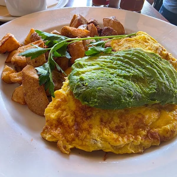 A diner breakfast plate with a golden omelet topped with sliced avocado, served with crispy home fries and parsley.