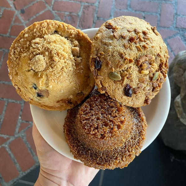 Hand holding a white plate with three muffins: blueberry streusel, bran, and a dark brown muffin.