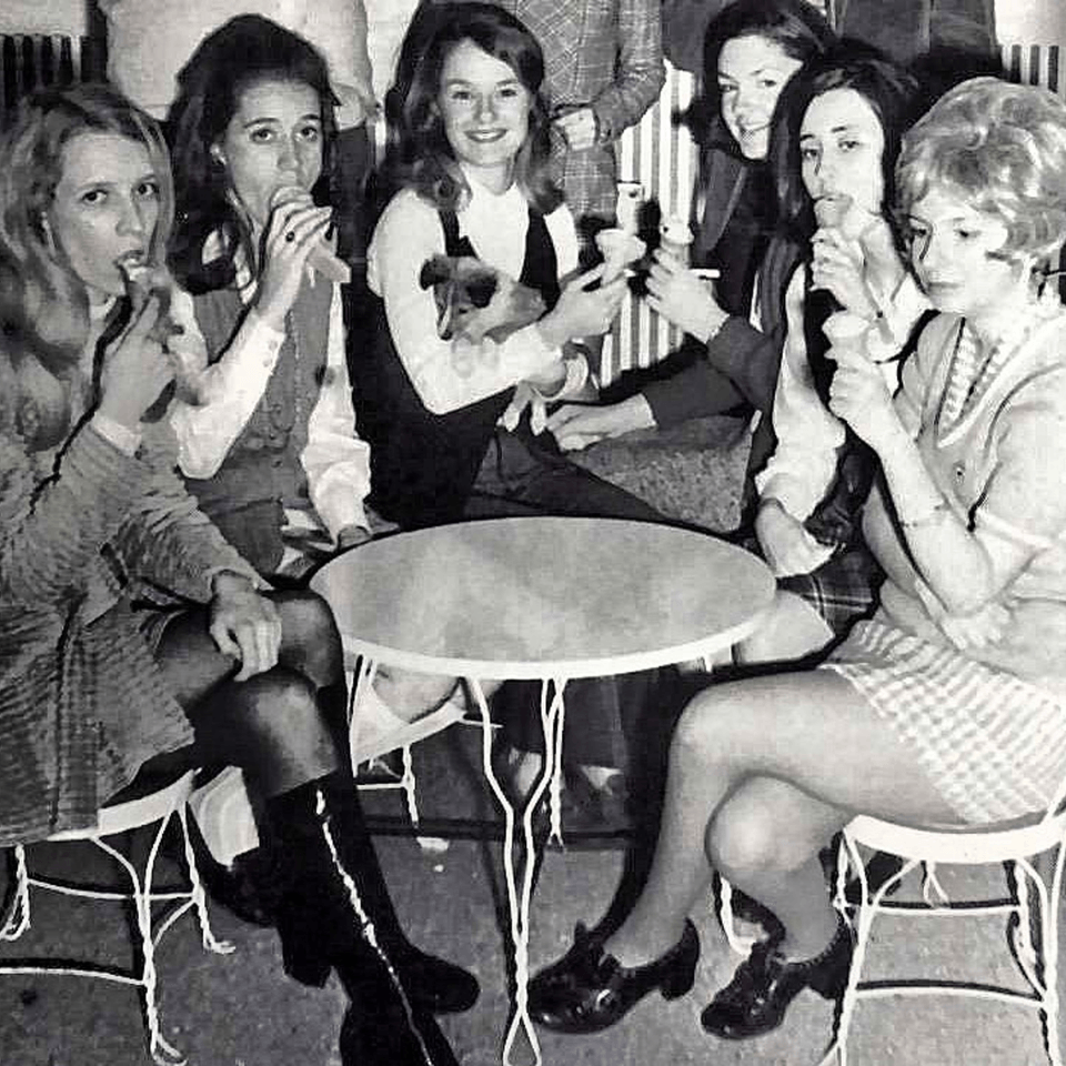 Six young women in 1960s attire enjoy ice cream cones around a small table at The Kettle diner.