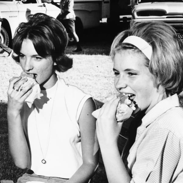 Two young women in 1960s attire eagerly eating sandwiches outdoors, with cars in the background.