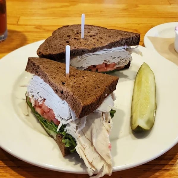 Turkey sandwich on dark bread with lettuce, tomato, and a pickle spear on a white plate.