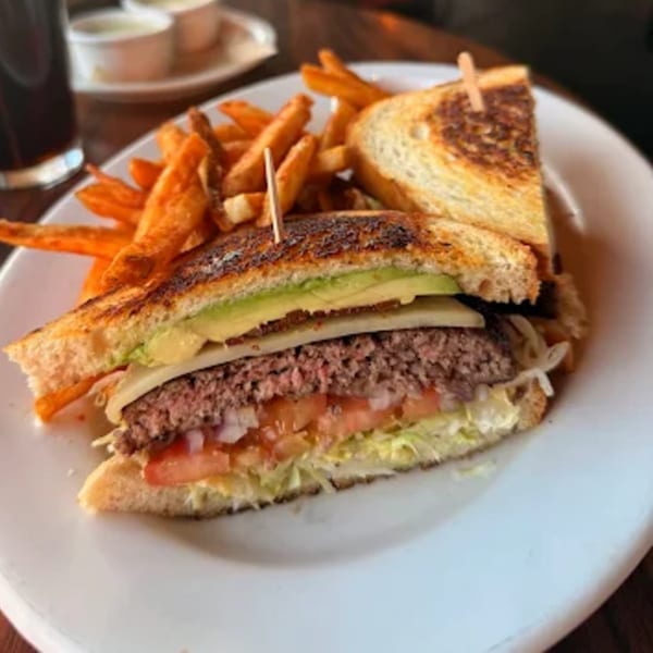 A grilled burger sandwich with avocado, cheese, lettuce, tomato, and fries on a white plate.