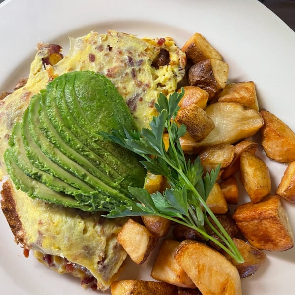 A breakfast plate with a fluffy omelet topped with sliced avocado and parsley, alongside crispy home fries.