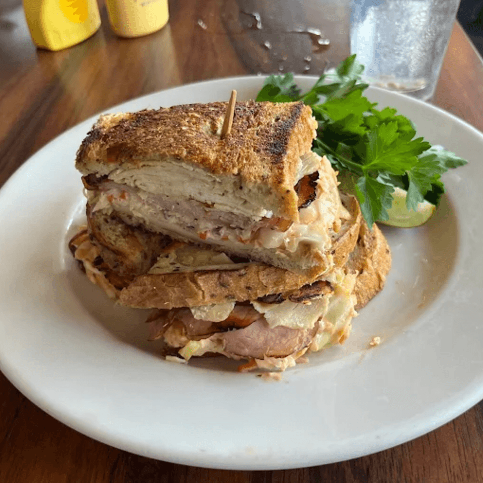A stacked deli sandwich with toasted bread, meat, and coleslaw, garnished with parsley and lime.