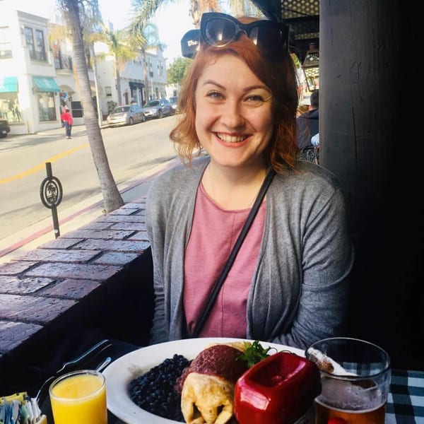 A smiling woman at The Kettle diner with a plate of food, orange juice, and beer on an outdoor patio.