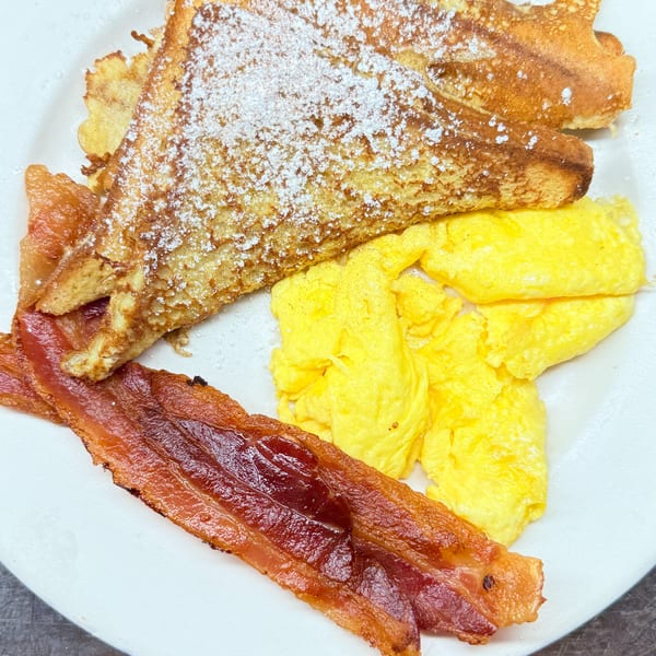 French toast with powdered sugar, scrambled eggs, and crispy bacon on a white plate.
