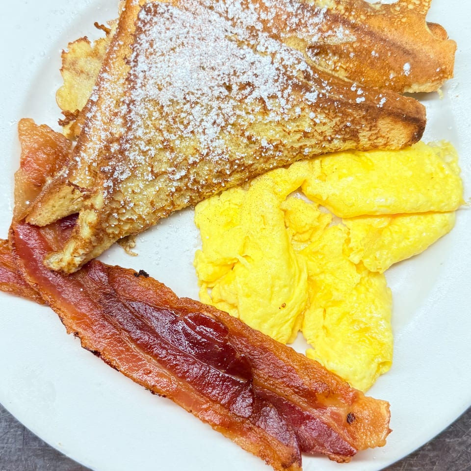 French toast with powdered sugar, scrambled eggs, and crispy bacon on a white plate.