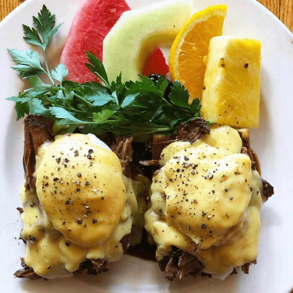 Eggs Benedict with hollandaise sauce and black pepper, served with a side of fresh fruit.